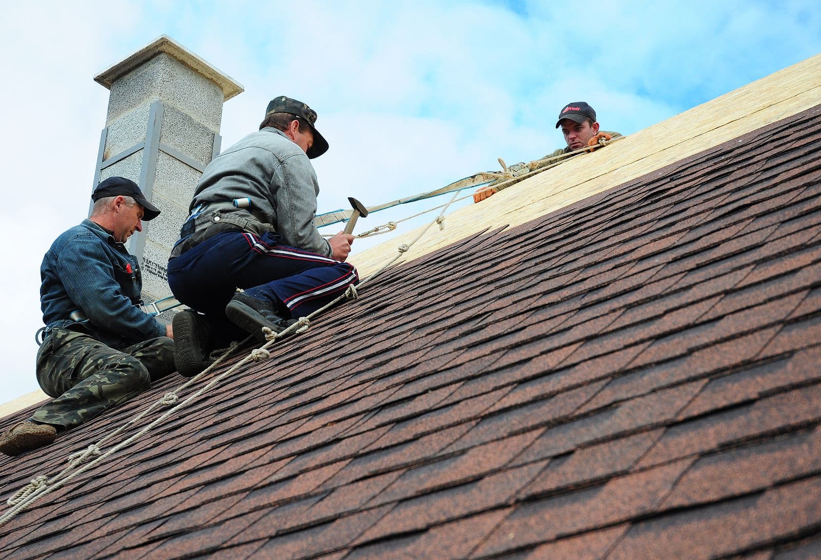 Three workers repair a steep, shingled roof. Two are kneeling with tools, while another observes from above. They are focused, ensuring safety.