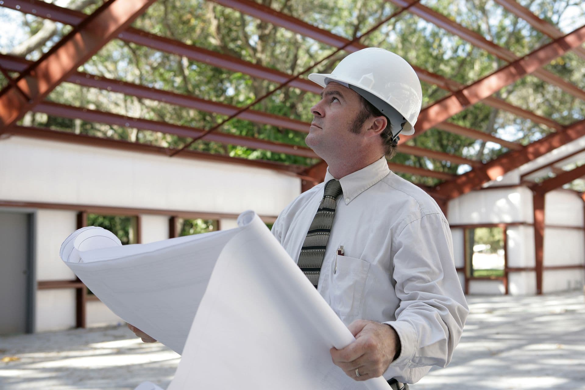A construction inspector holding blueprints and looking at the roof beams of a steel building in progress.