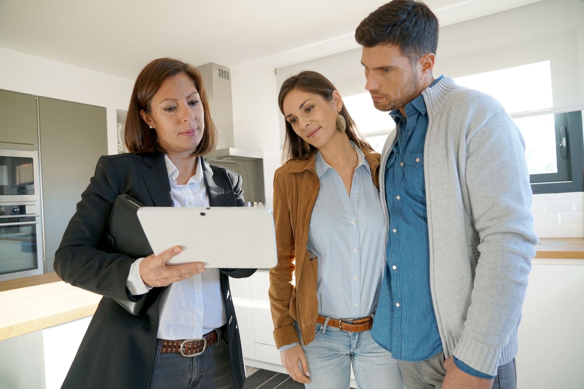 An agent shows a document on a clipboard to a couple in a bright, modern kitchen. The couple looks attentively, suggesting decision-making.