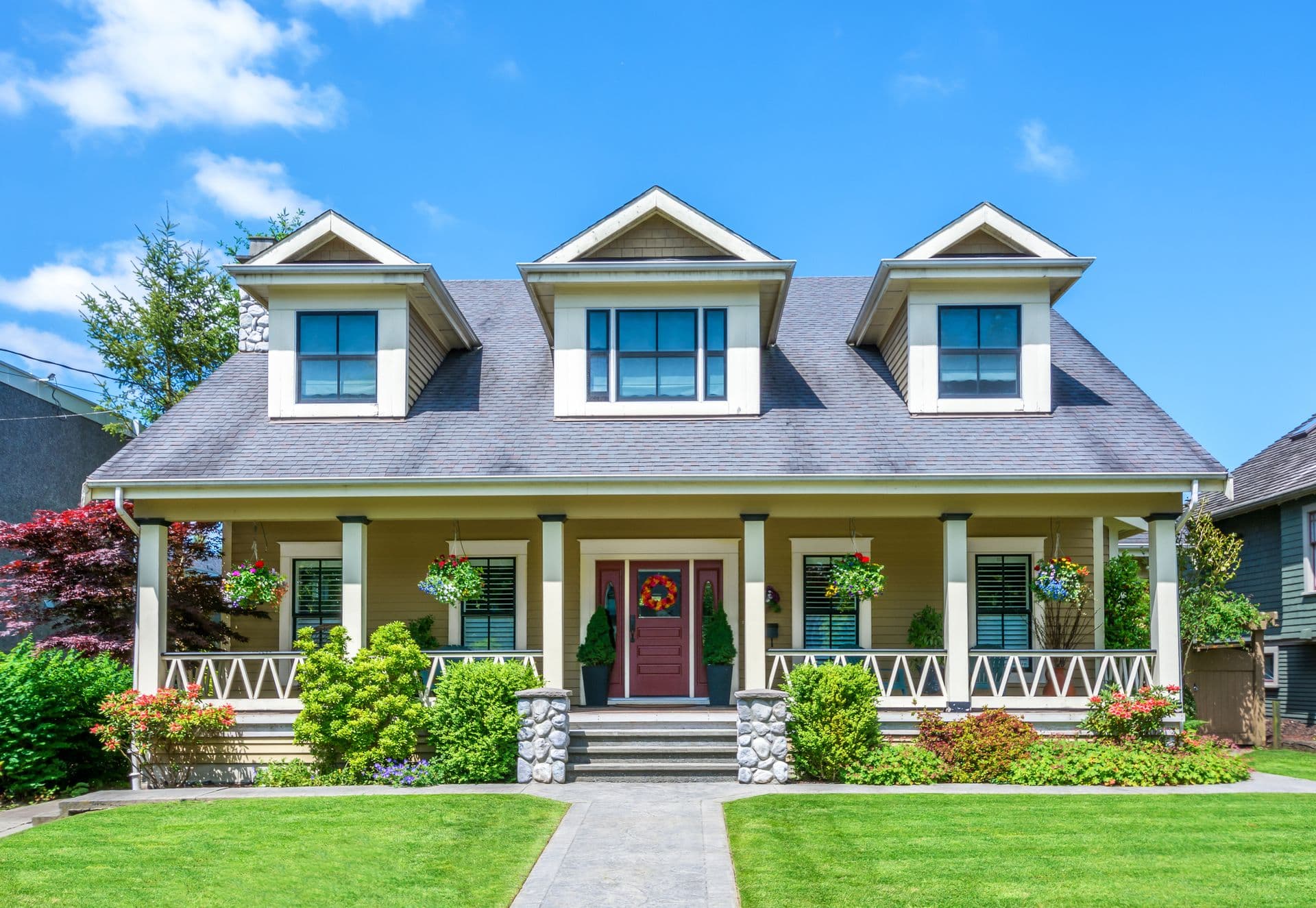 A charming two-story craftsman house with dormer windows, a red front door, and a porch adorned with hanging plants. Lush green lawn and vibrant shrubs.
