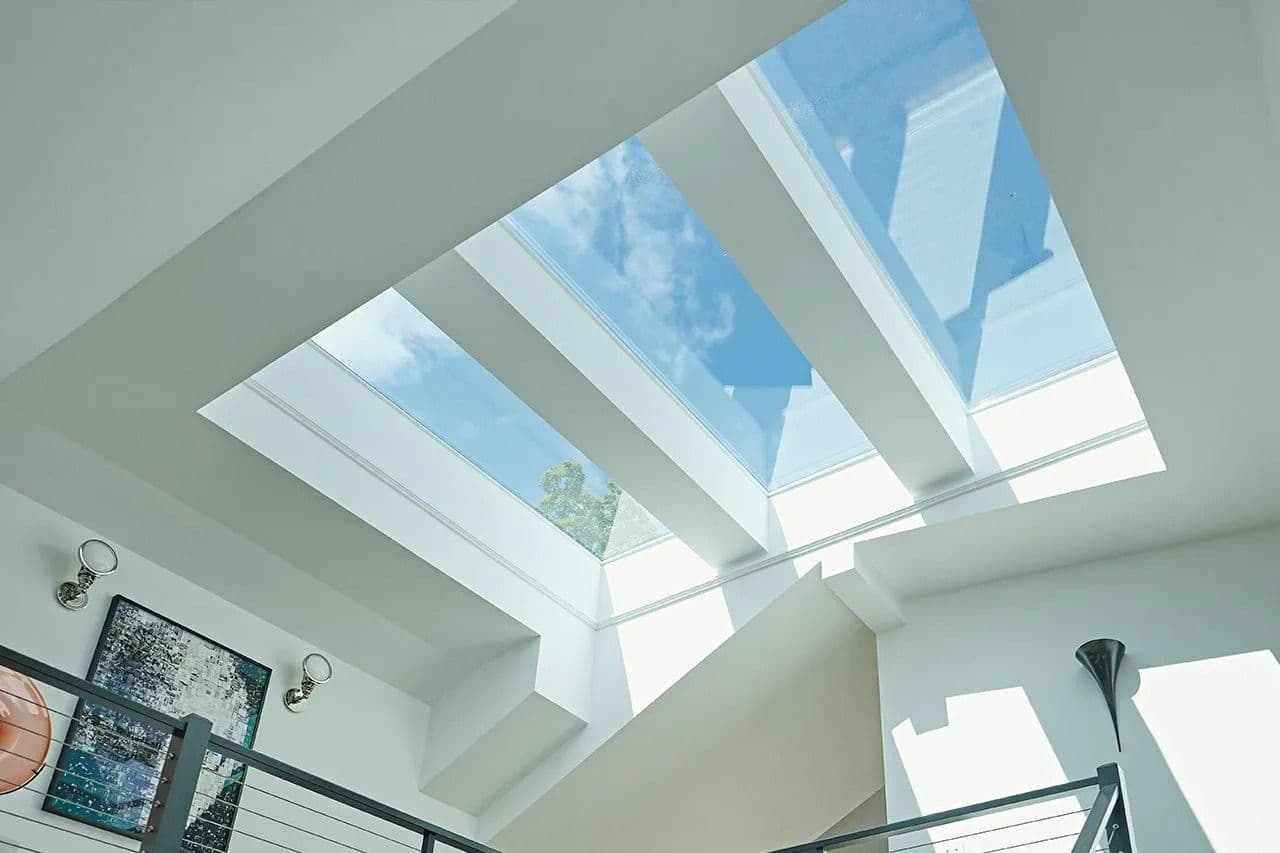 Modern interior with a large skylight ceiling featuring four glass panels, allowing blue sky and clouds to be visible. White walls create a bright, airy feel.