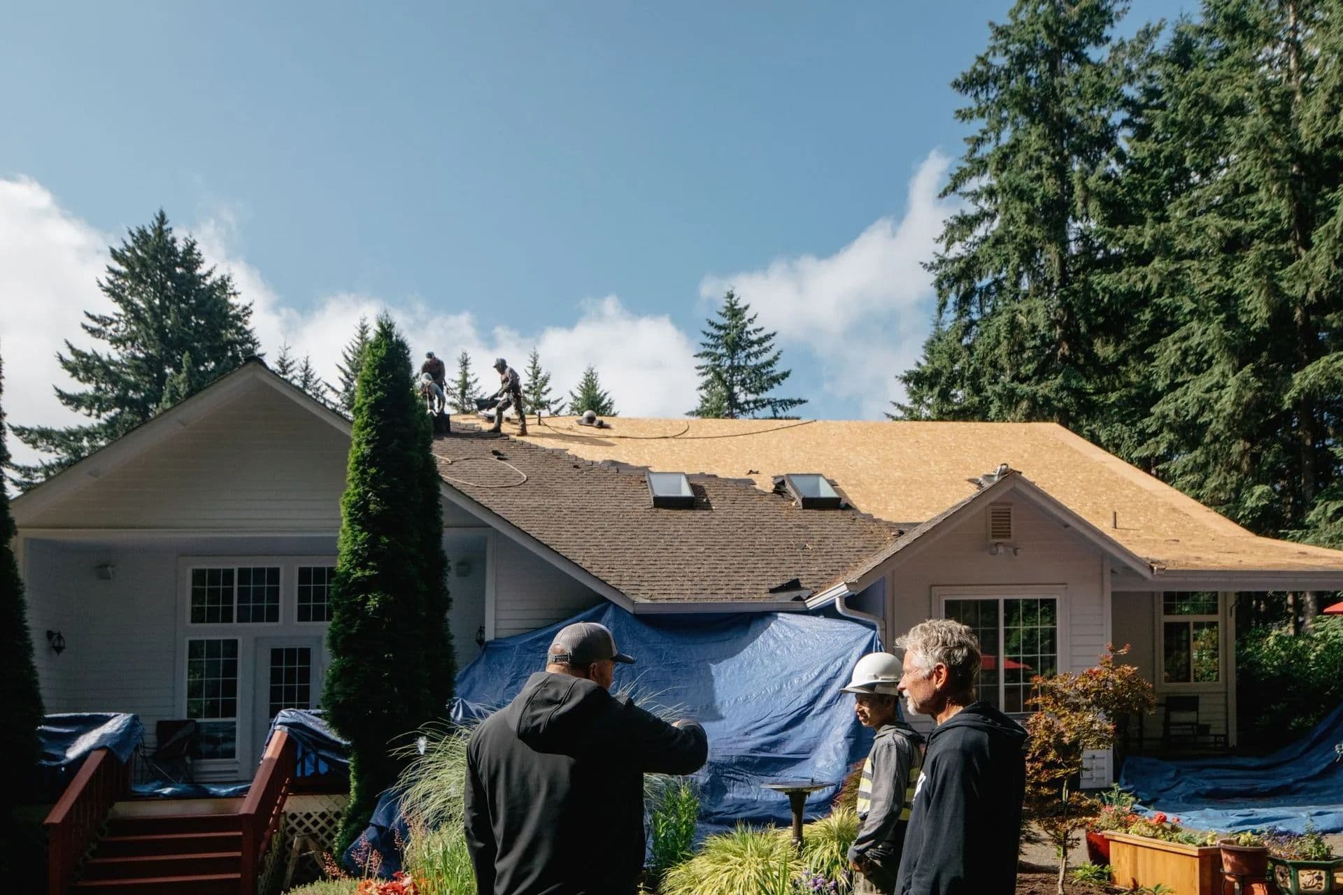 A group of workers on a partially completed house roof surrounded by tarps. Tall trees in the background. People on the ground observe the progress.