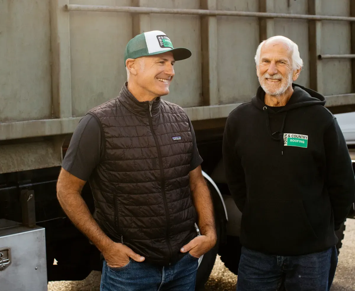 Two men stand smiling in front of a large truck. One wears a quilted vest and cap, the other a hoodie. They appear friendly and relaxed.