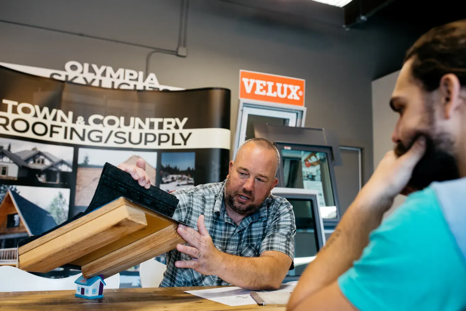 A man in a plaid shirt explains a roofing sample to a seated customer in a roofing supply showroom. The setting is professional and informative.
